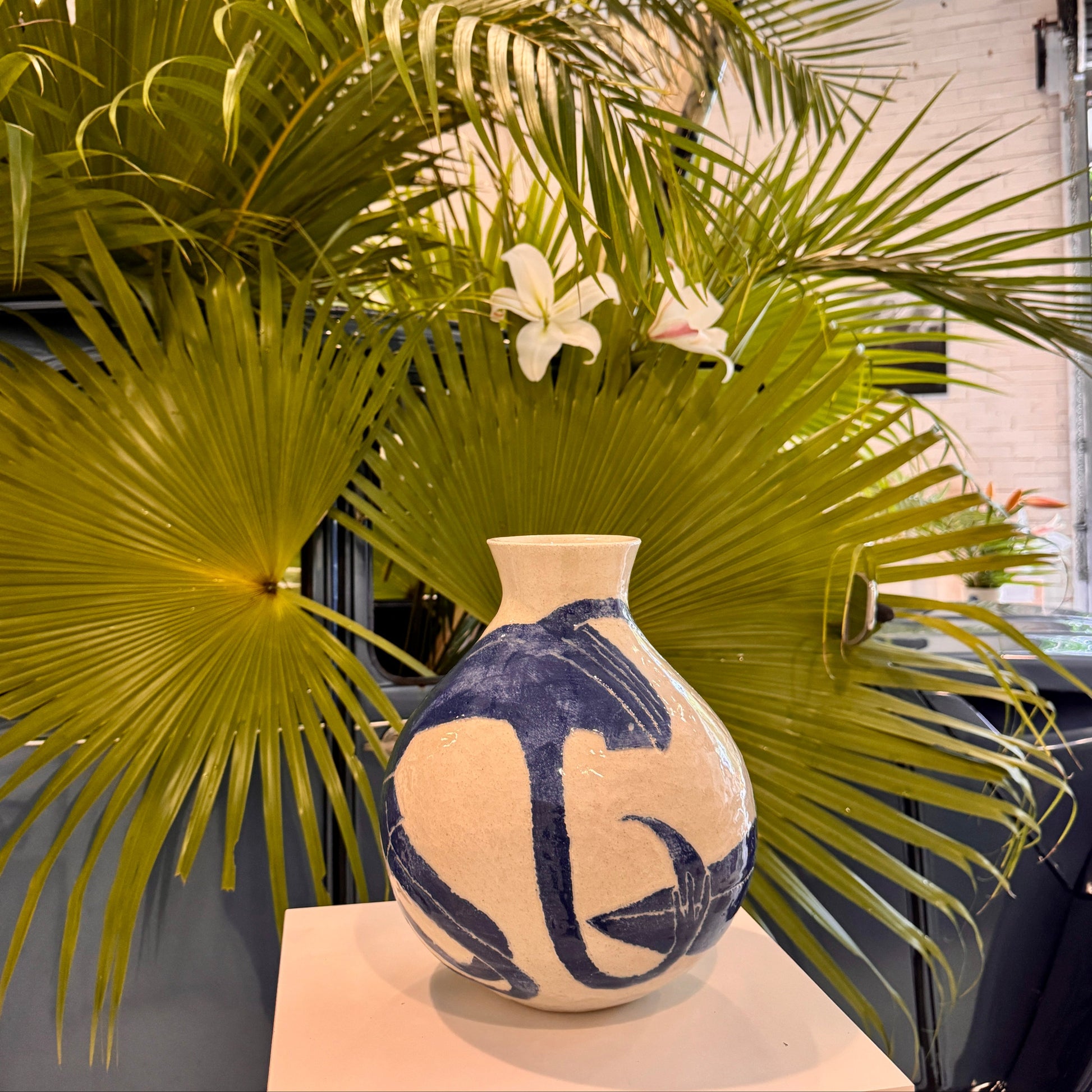 Decorative vase by Damion Fuller with blue and white patterns on a surface with green palm leaves in the background.