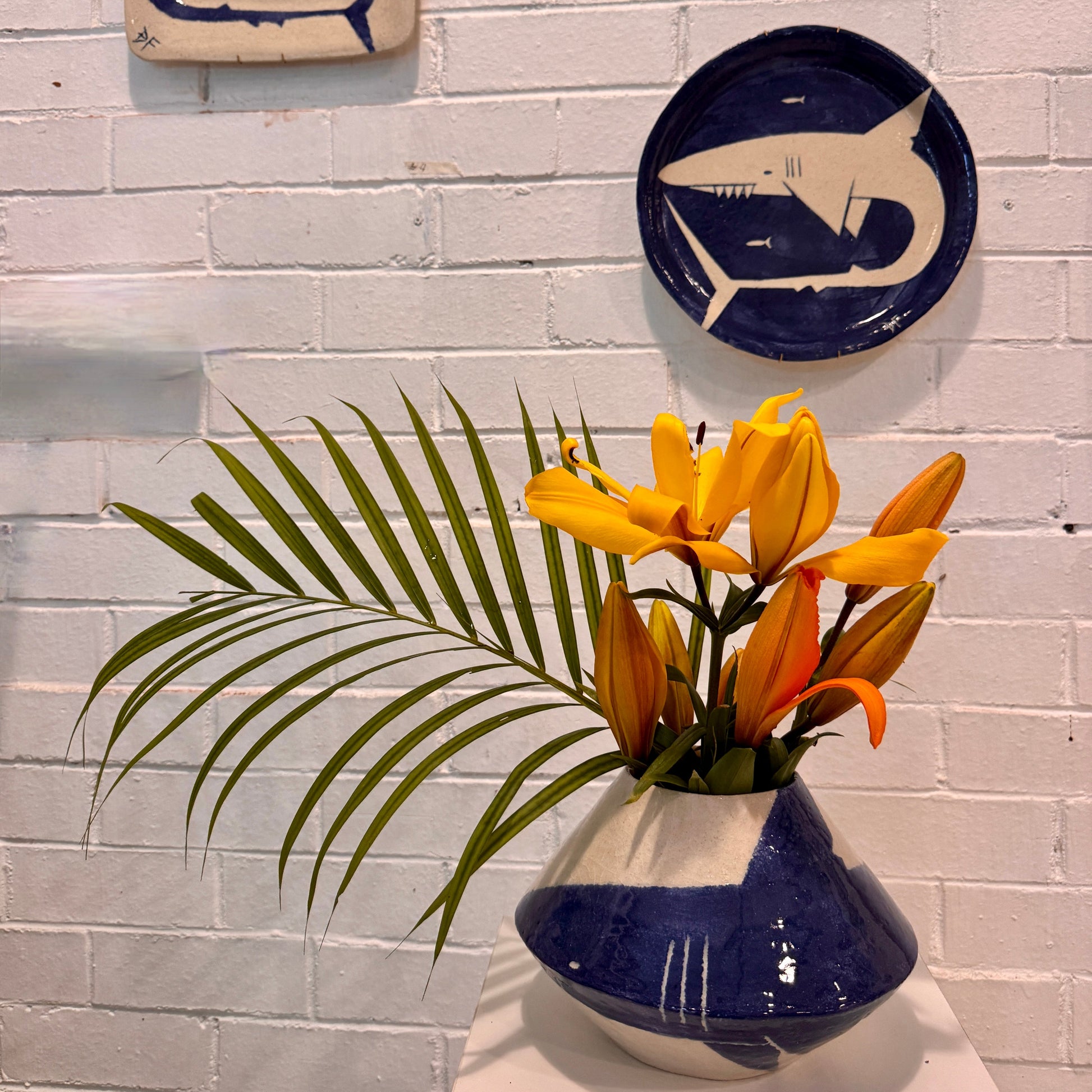 Decorative plates with shark designs by Damion Fuller on a white brick wall, accompanied by a vase with flowers.