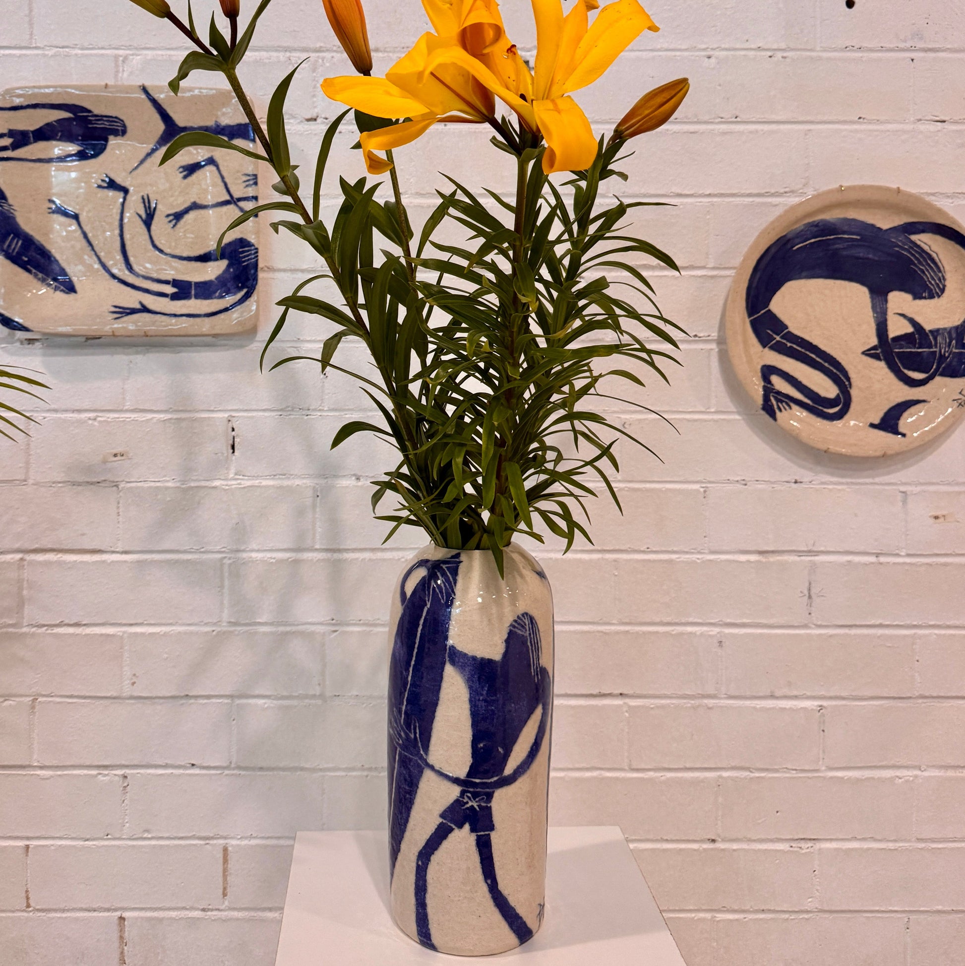 Vase by Damion Fuller with yellow flowers on a white surface against a light gray brick wall with decorative plates.