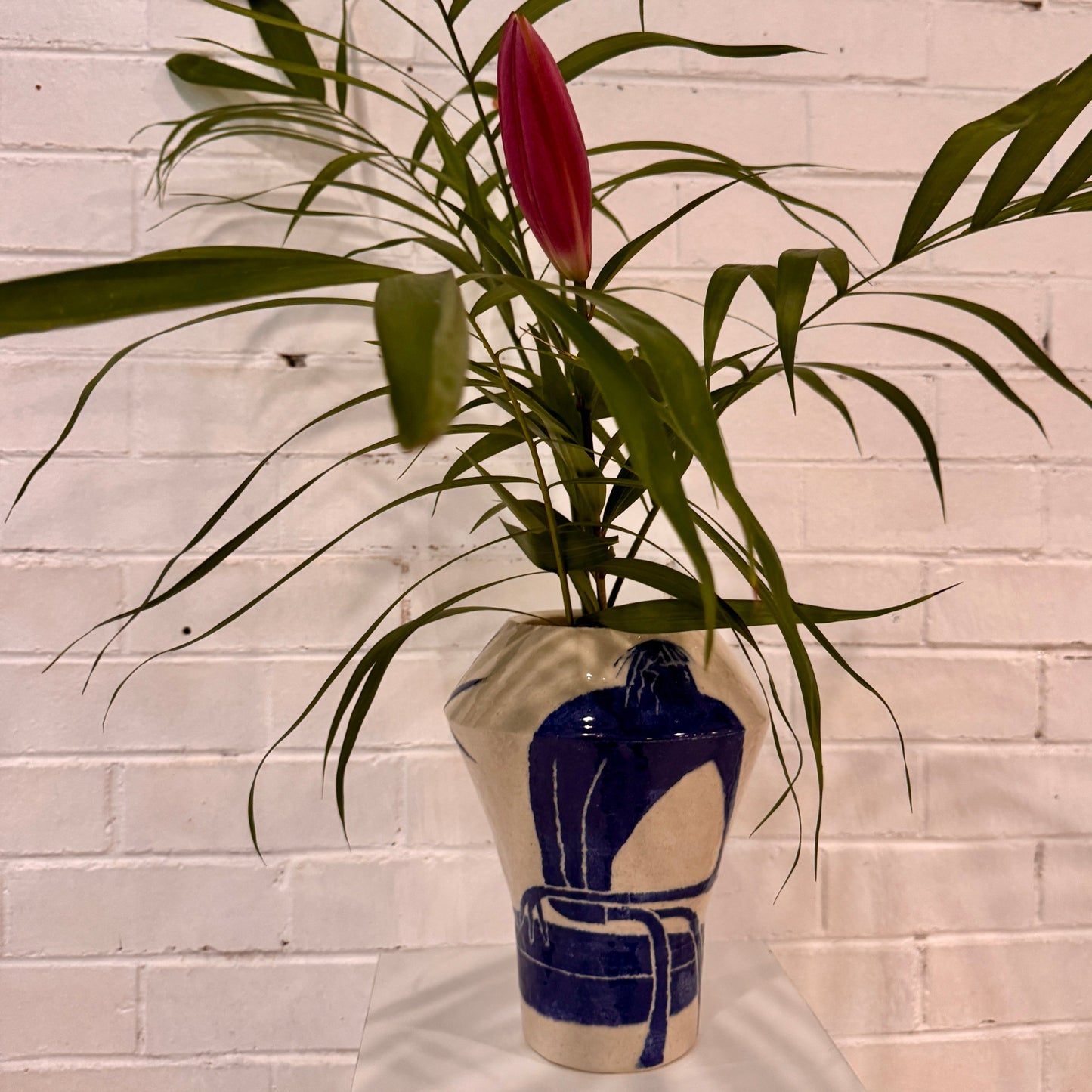 Vase by Damion Fuller with green leaves and a red flower against a white brick wall